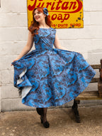 A woman with red hair wearing a blue and gray patterned 1950s-style swing dress, posing on a wooden bench in front of a white brick wall with a vintage yellow soap sign.