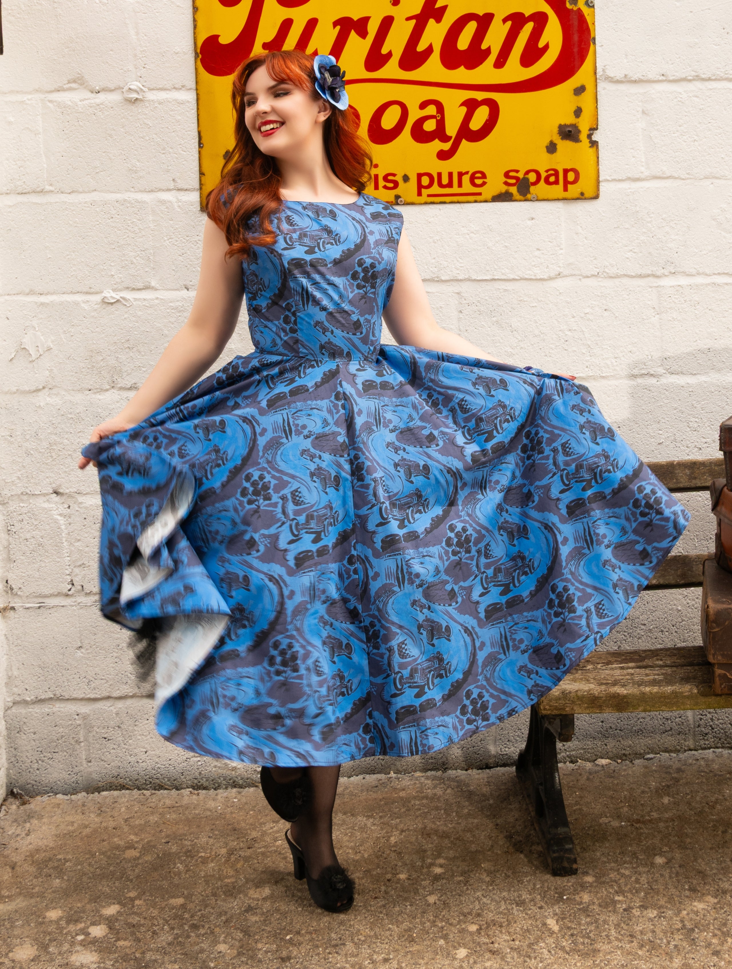 A woman with red hair wearing a blue and gray patterned 1950s-style swing dress, posing on a wooden bench in front of a white brick wall with a vintage yellow soap sign.
