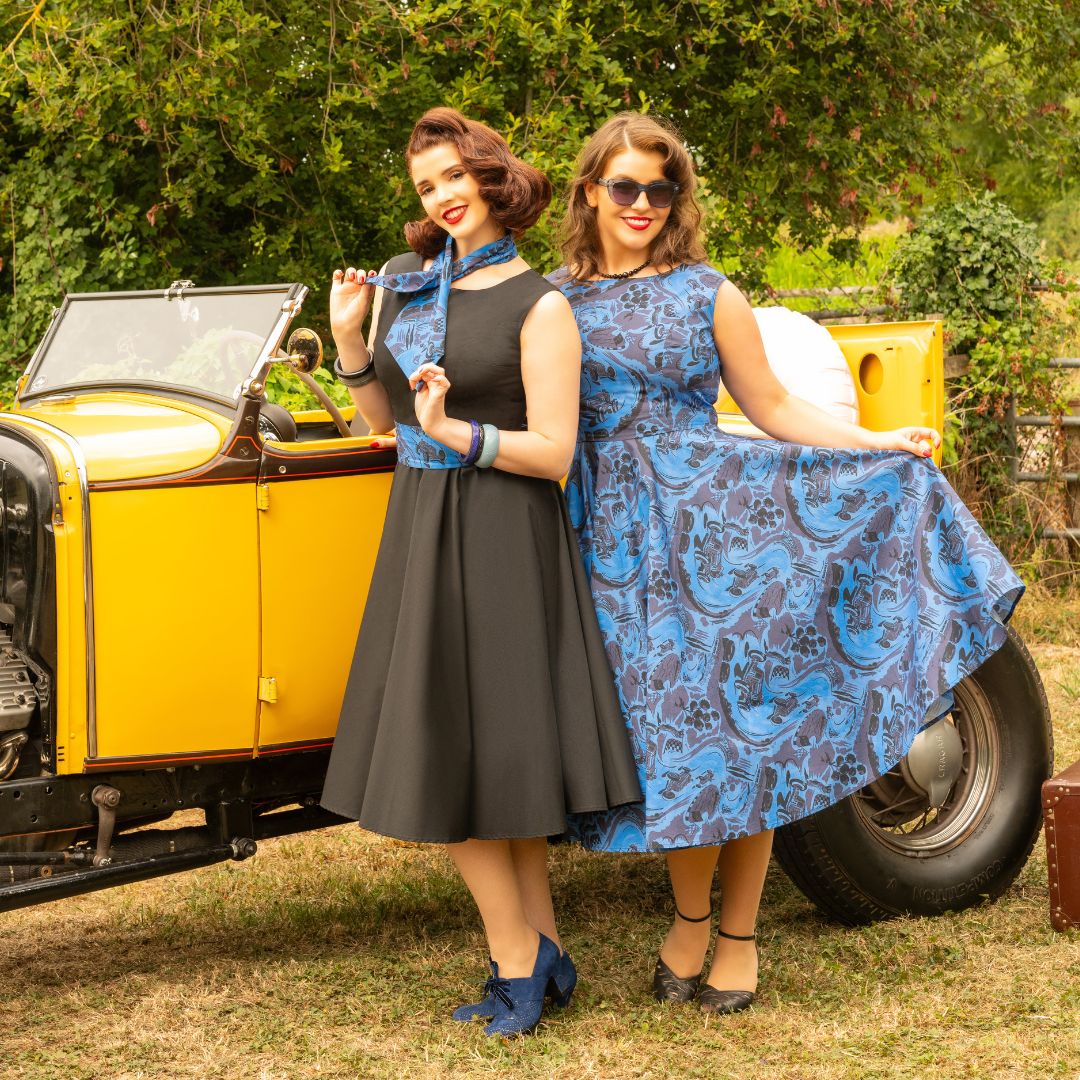 Two women posing outdoors in front of a vintage yellow car. The woman on the left wears a black sleeveless dress with a blue patterned neck scarf and blue shoes.