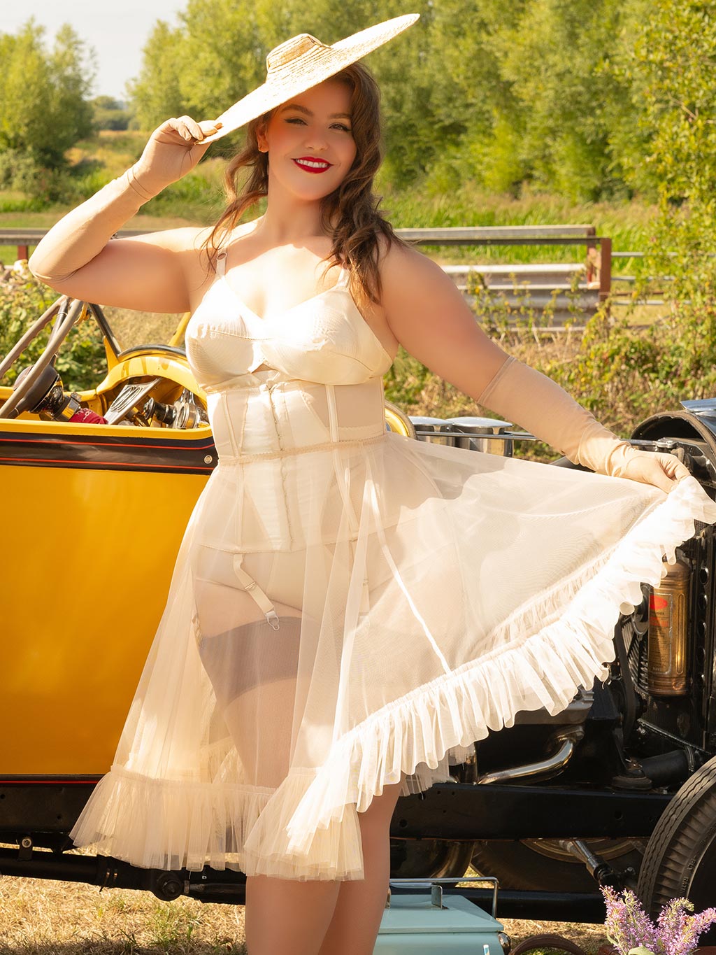 Woman in vintage lingerie and petticoat and hat standing next to a yellow car in a field.