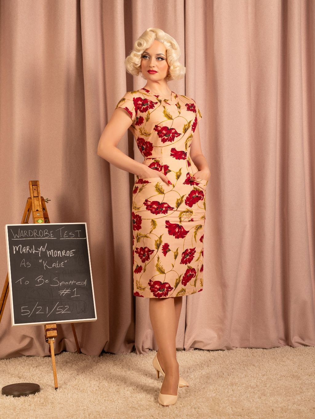 Woman in a floral dress standing next to a blackboard with text, against a pink curtain background.