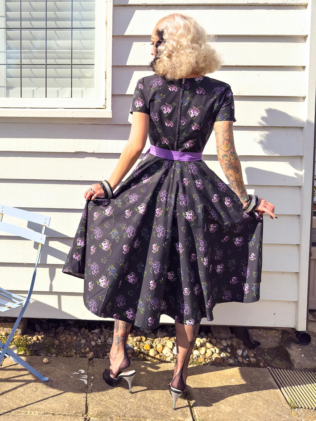 back view of a 1950s purple floral dress with a square neckline and short sleeves worn with black seamed stockings