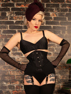 A woman poses in a vintage-style black lingerie set featuring a corseted bustier, matching garter belt, sheer long gloves, and thigh-high stockings, paired with a dark beret, set against a brick wall background.