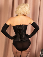 Person wearing a black corset and fishnet stockings against a beige curtain.