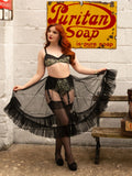 A woman with red hair models a vintage-style lingerie set featuring a black and olive green floral patterned bra and garter belt, a sheer black fringed tutu skirt, and black thigh-high stockings, posed in front of a brick wall with a retro Puritan Soap sign.