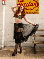 A woman posing in a black lace and sheer tulle costume with garter details, standing in front of a white brick wall with a vintage yellow Suritan Soap sign and a wooden bench.