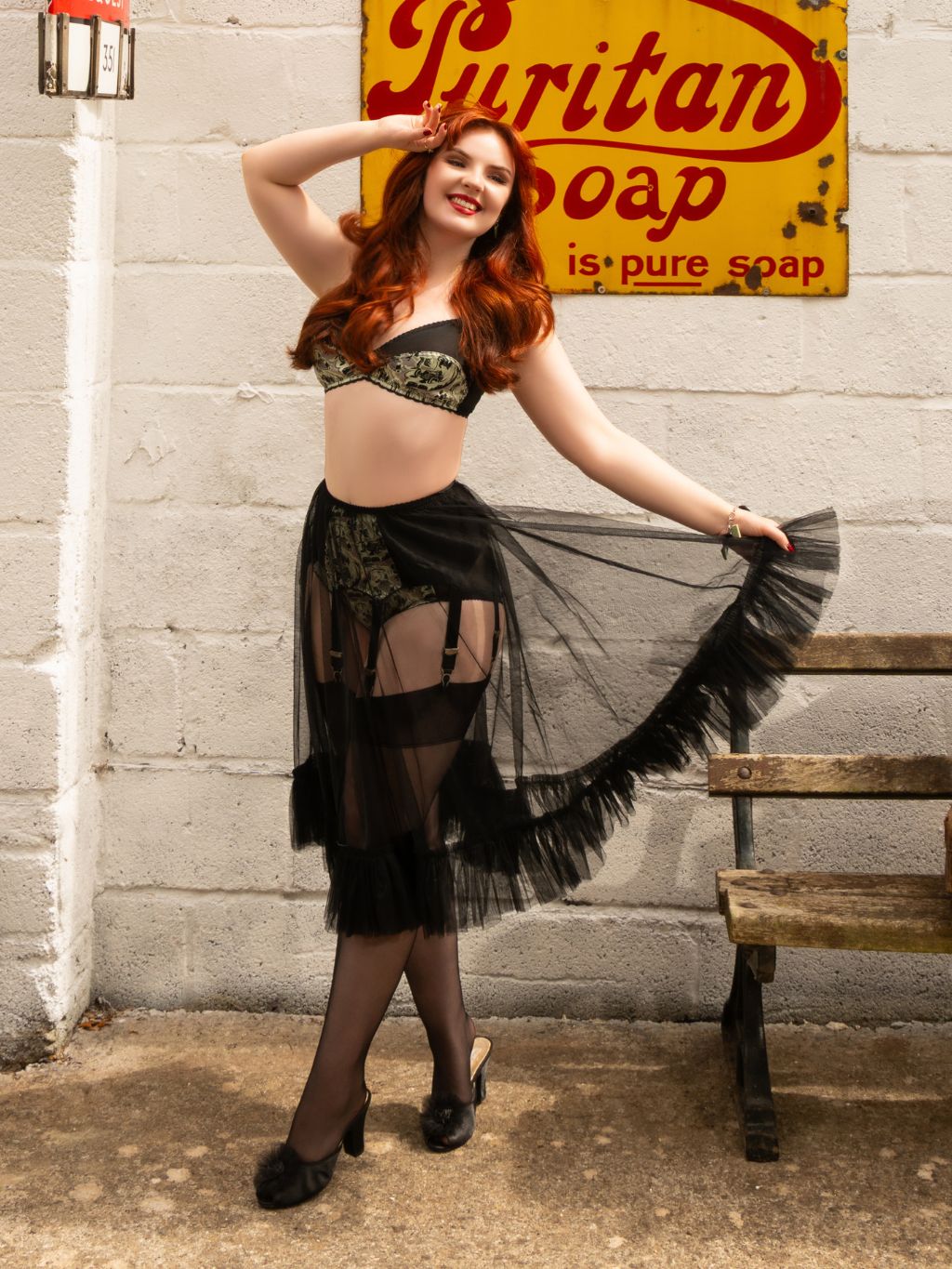 A woman posing in a black lace and sheer tulle costume with garter details, standing in front of a white brick wall with a vintage yellow Suritan Soap sign and a wooden bench.