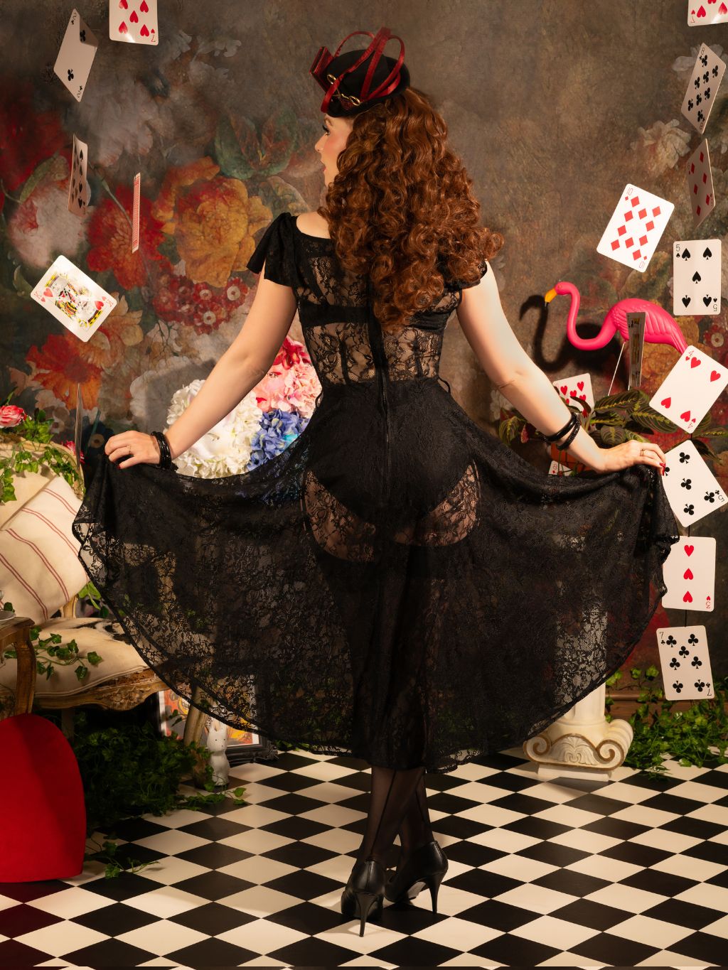 Woman in a black lace dress standing in a room with playing cards and flamingos.