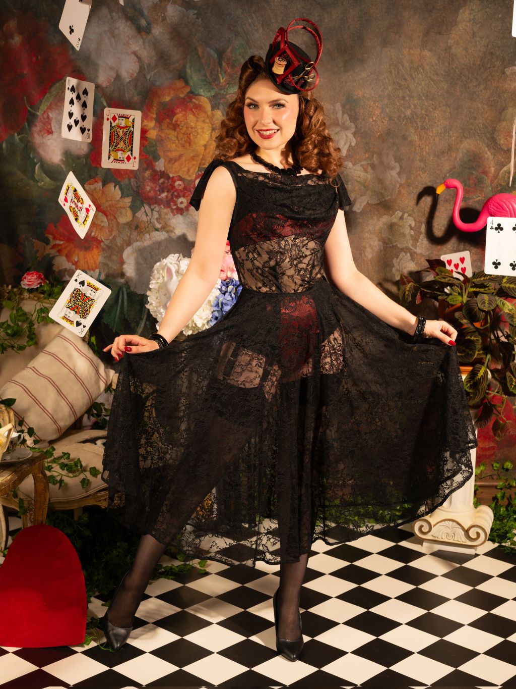 Woman in a black lace dress posing in a room with playing cards and decorative elements.