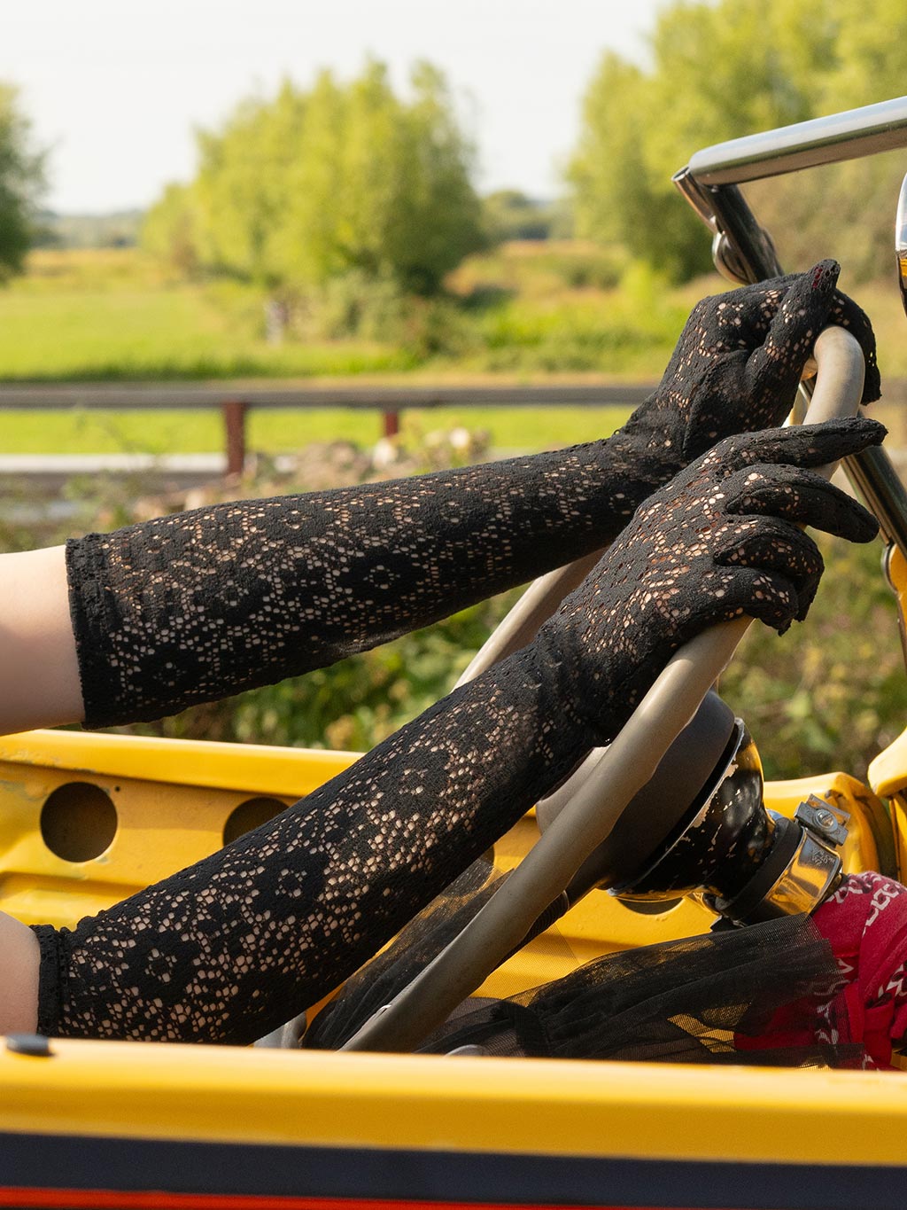 Person wearing black lace elbow length gloved, steering a vintage car with greenery in the background