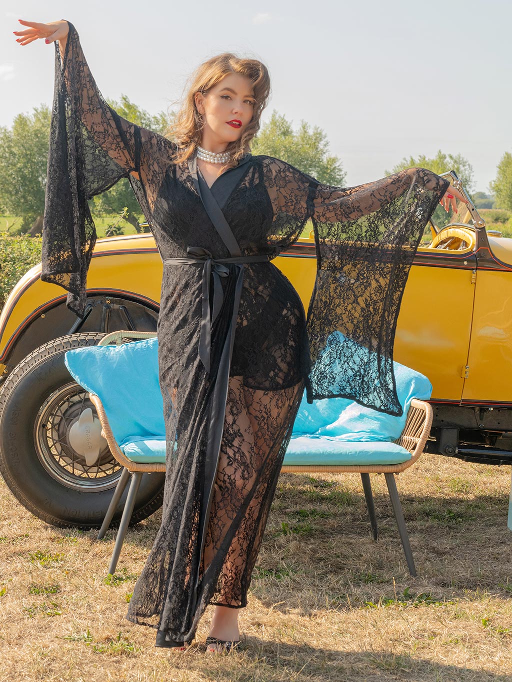 Woman in a black lace kimono standing in front of a vintage car with a blue chair.