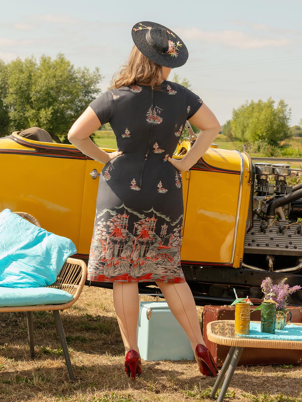 Woman in a vintage dress standing outdoors with a yellow vehicle and table in the background