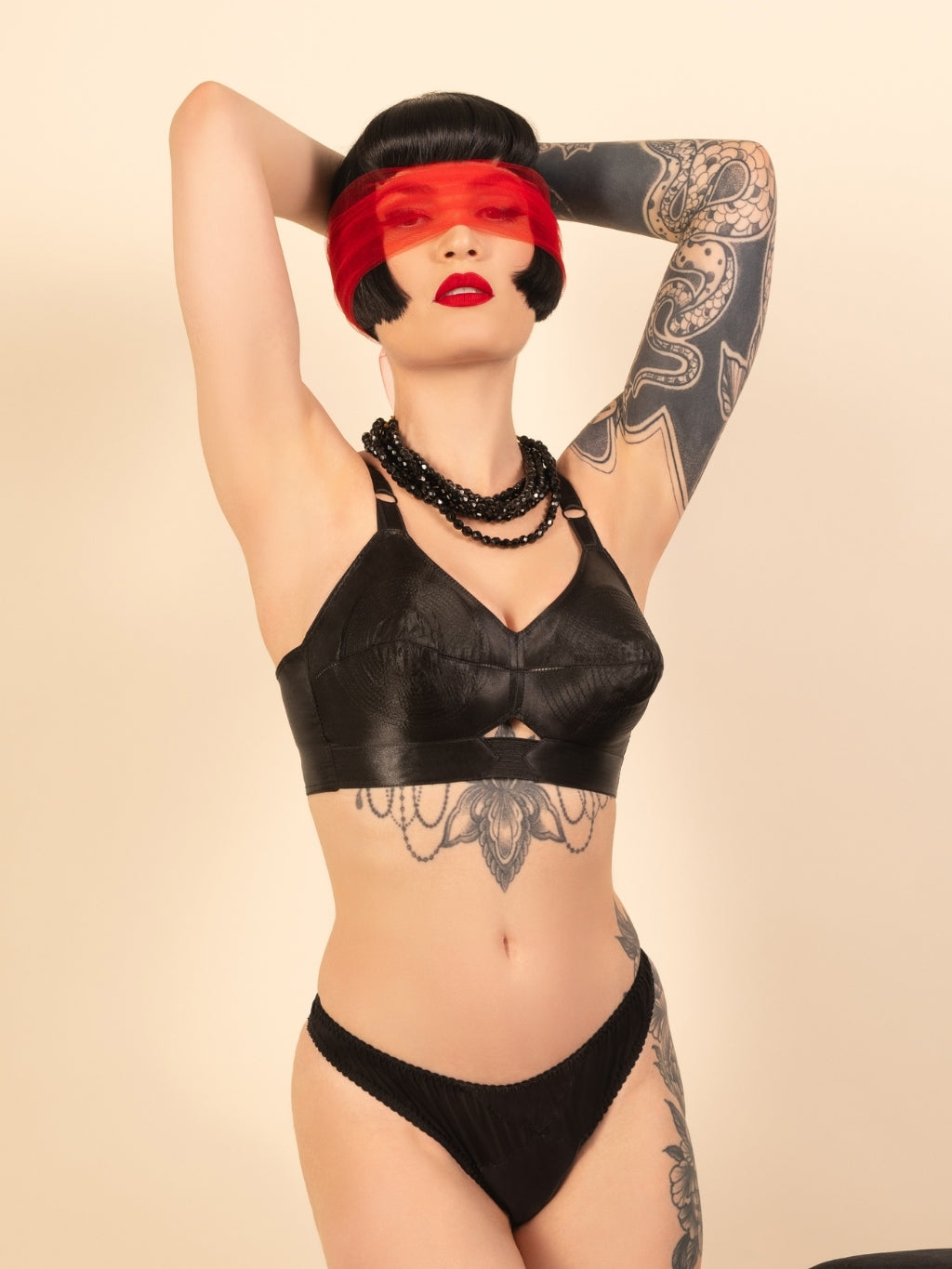 Model wearing a black satin bullet bra and thong set with tattoos on arms and chest against a beige background.