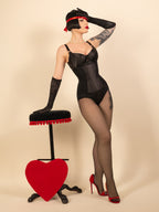 Woman in black lingerie and black seamed tights posing with a red heart-shaped stool on a beige background