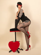 Woman in black lingerie and seamed tights sitting on a red heart-shaped stool against a beige background