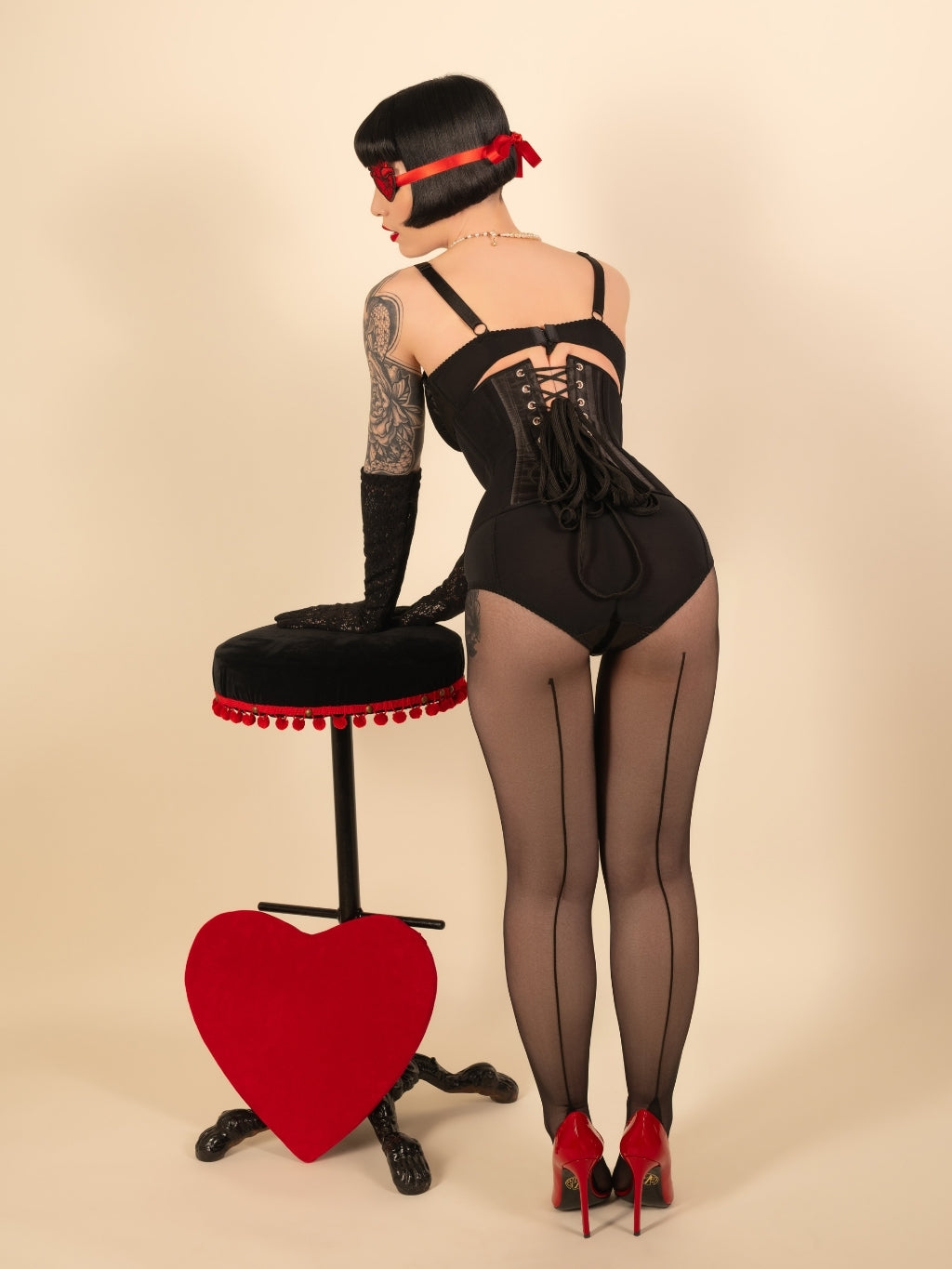 Woman in black lingerie and seamed tights standing next to a red heart-shaped object on a beige background