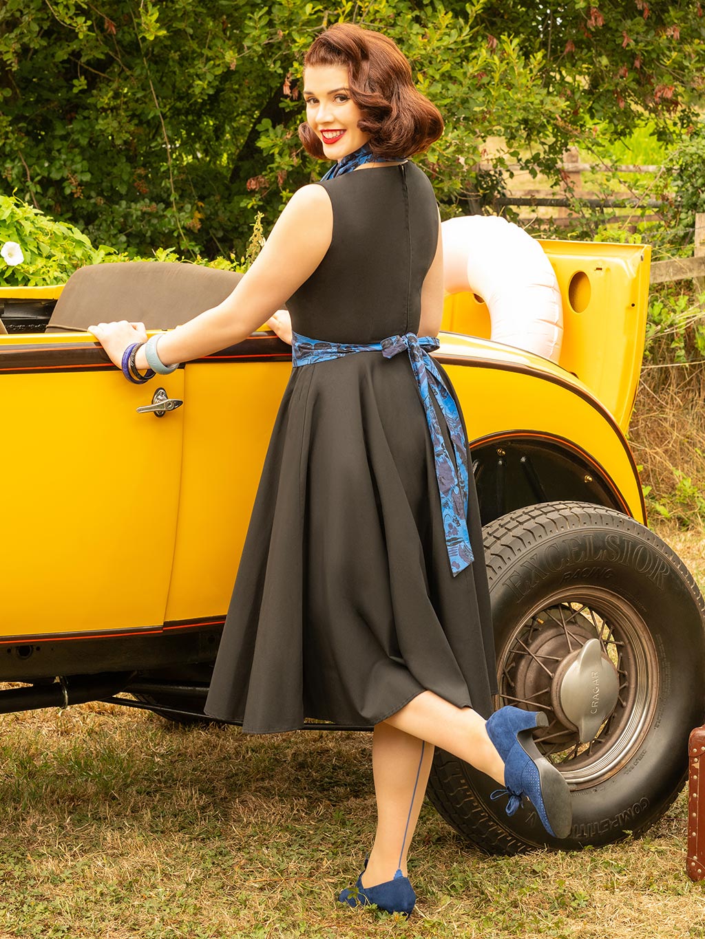 Woman in a black dress with a blue apron standing next to a yellow vintage car.