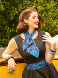Woman in a vintage-style dress with a blue scarf, sitting in a yellow car with greenery in the background.