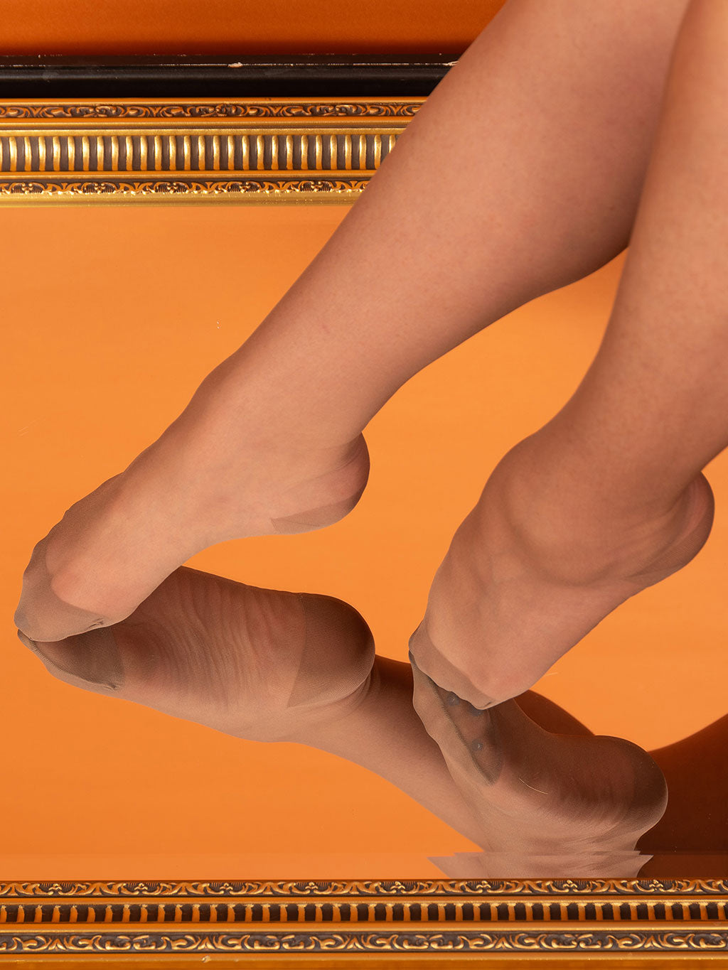Close-up of legs wearing sheer brown rht stockings with a reflective surface and gold frame in the background.