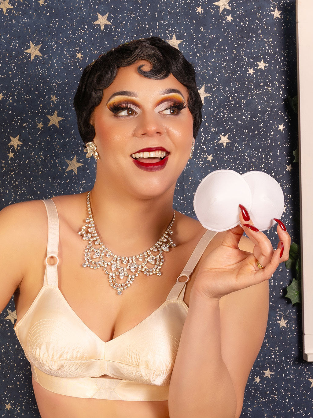 Woman in a peach satin bullet bra holding white bullet bra pads against a starry background