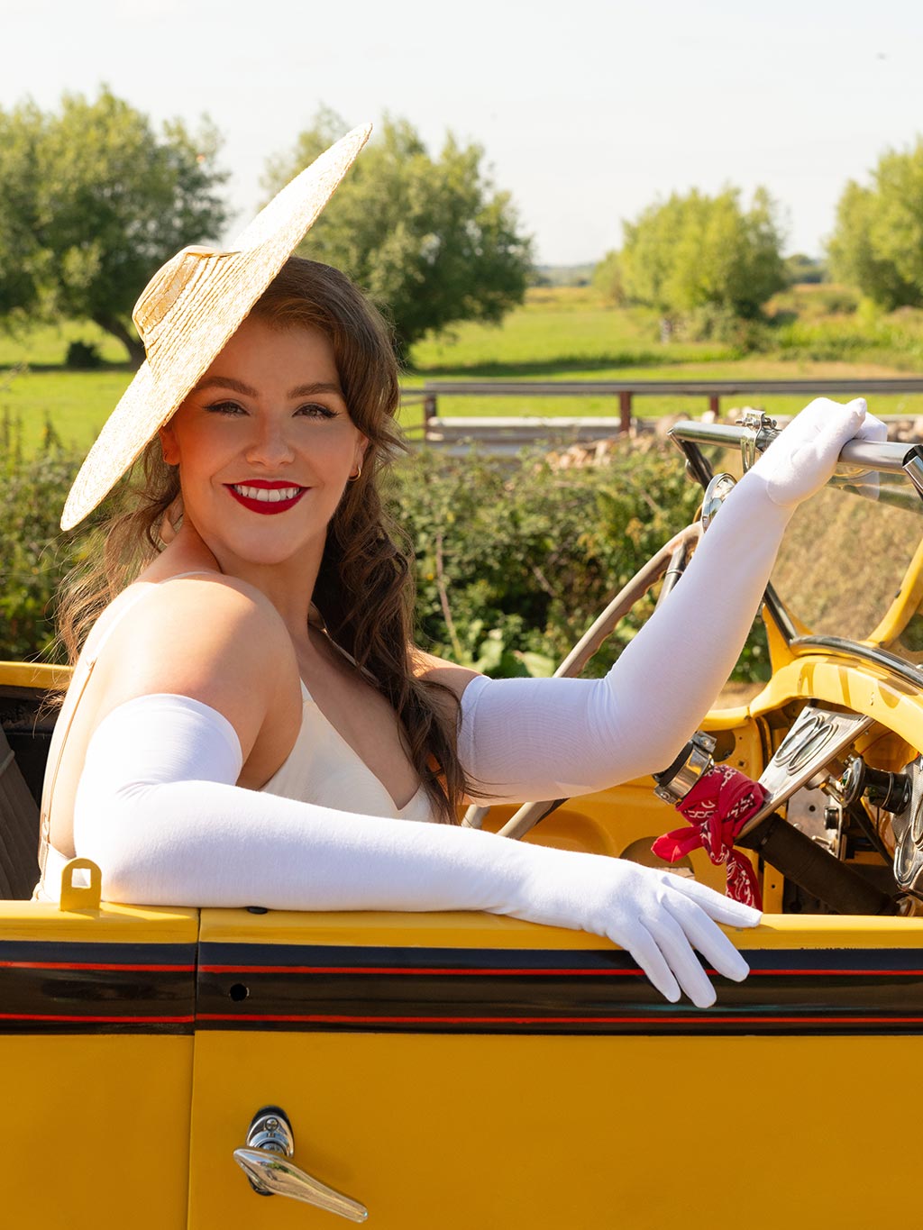 Woman in a vintage car wearing a wide-brimmed hat and white gloves, with a scenic background.