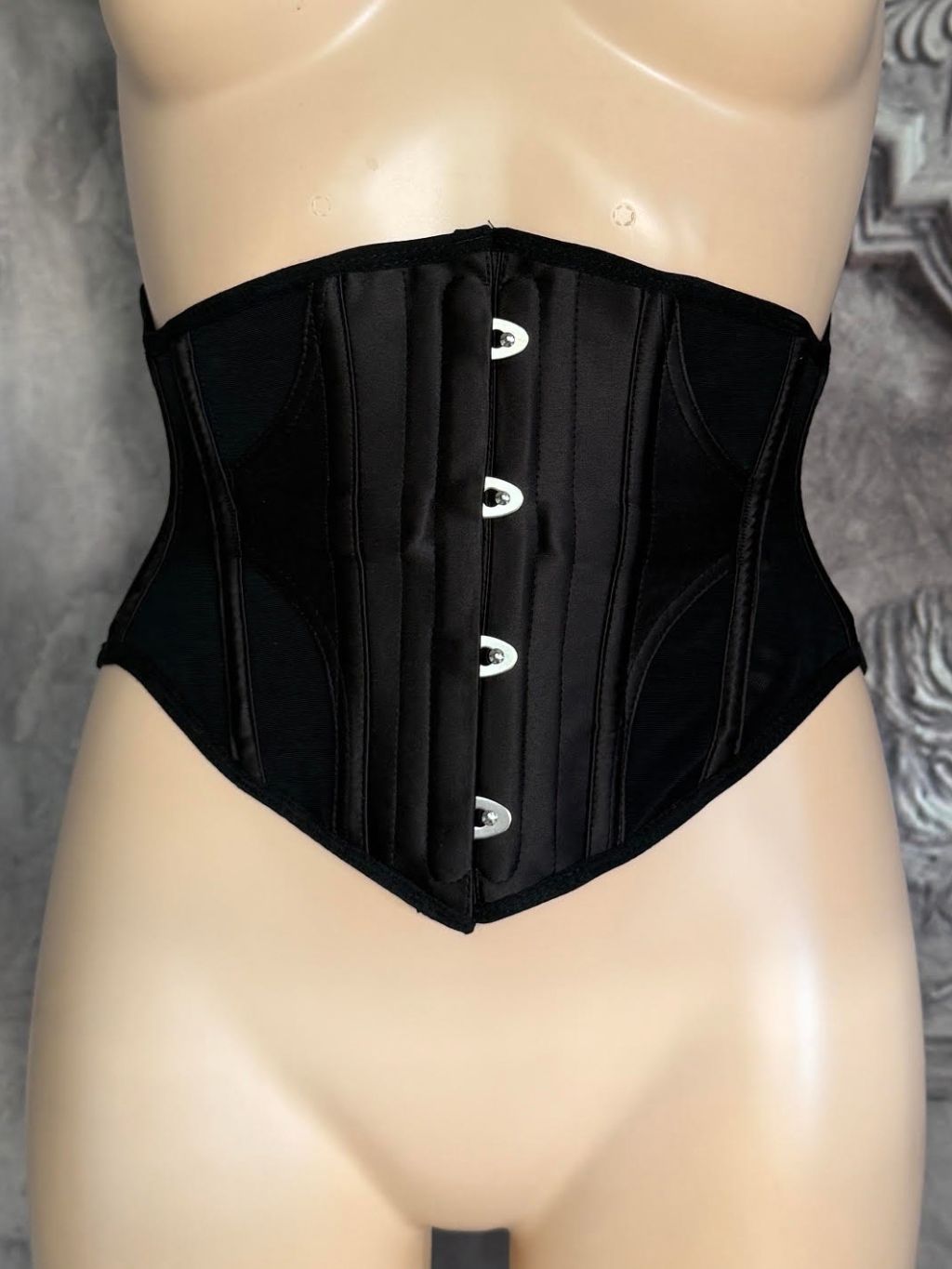 A black structured waist trainer corset displayed on a beige mannequin torso against a gray textured background.