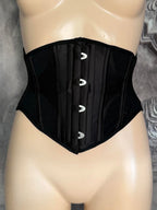 A black structured waist trainer corset displayed on a beige mannequin torso against a gray textured background.