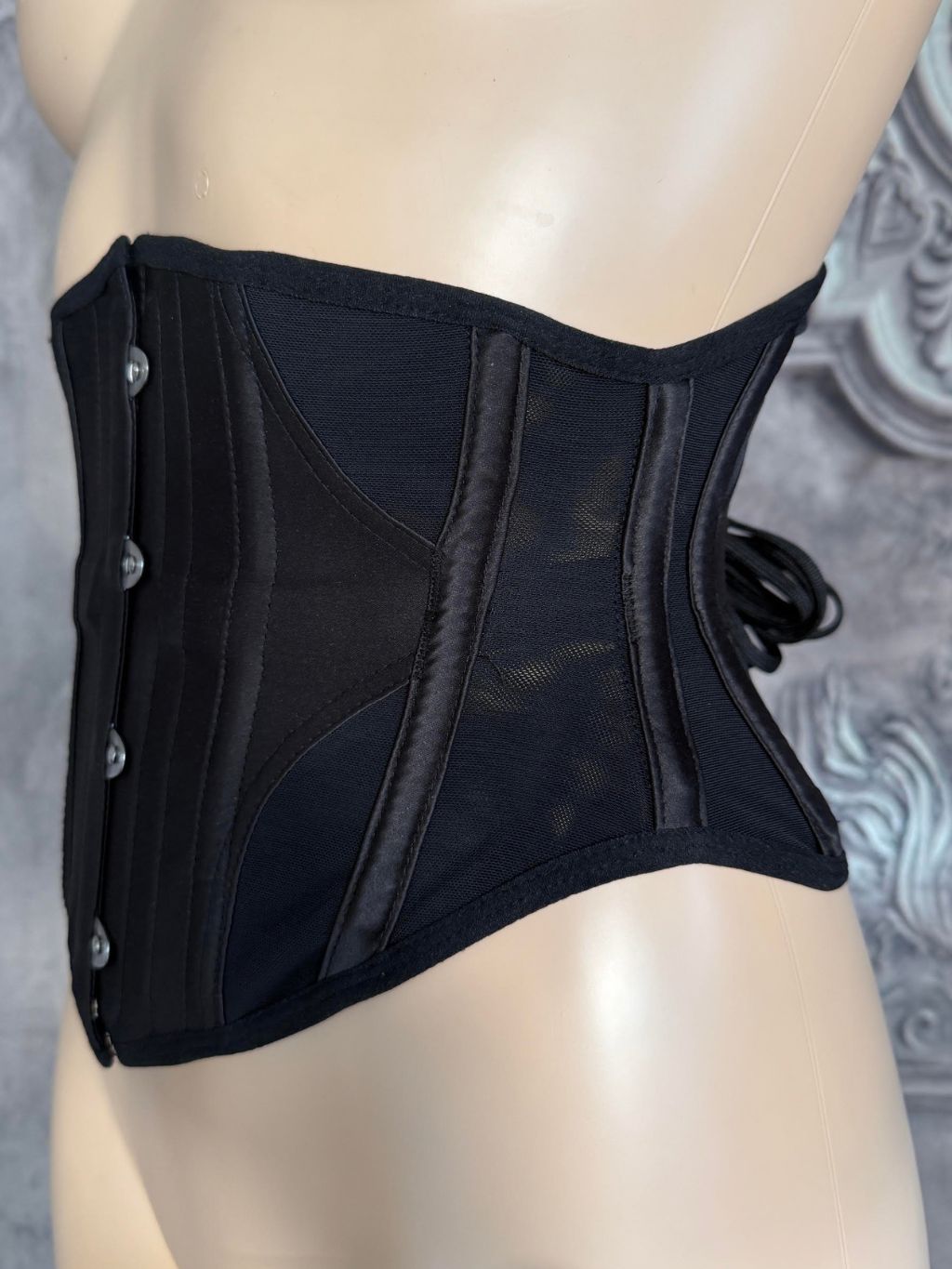 A black structured corset waist trainer with front buttons and side laces, displayed on a beige mannequin torso against a gray patterned backdrop.
