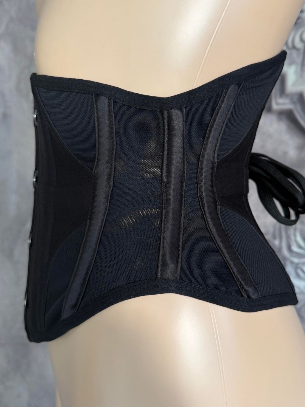 A black structured waist trainer corset with sheer mesh panels and side laces, displayed on a beige mannequin torso against a gray patterned background.