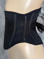 A black structured waist trainer corset with sheer mesh panels and side laces, displayed on a beige mannequin torso against a gray patterned background.