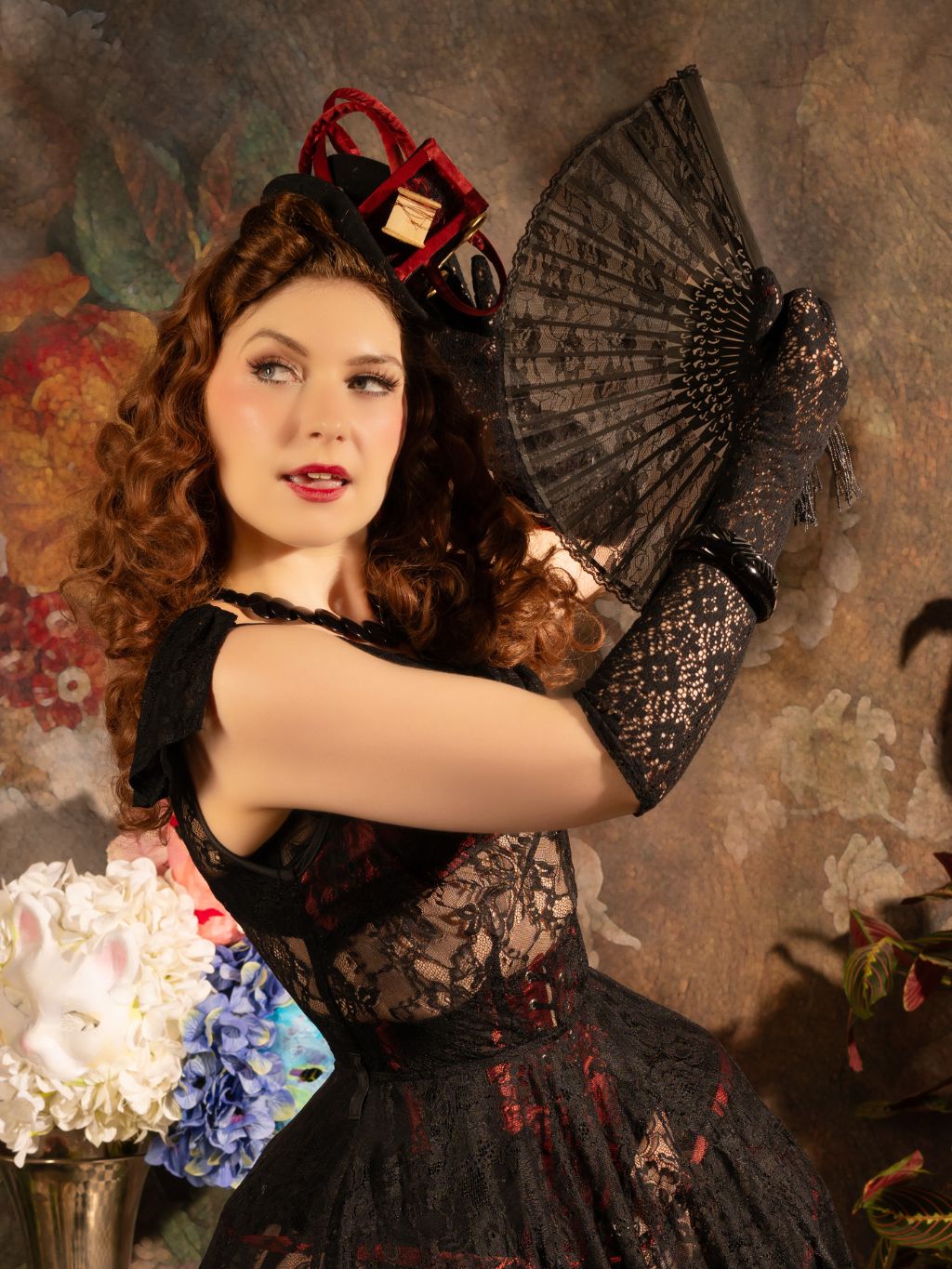 Woman in a black lace dress and gloves holding a fan against a decorative background