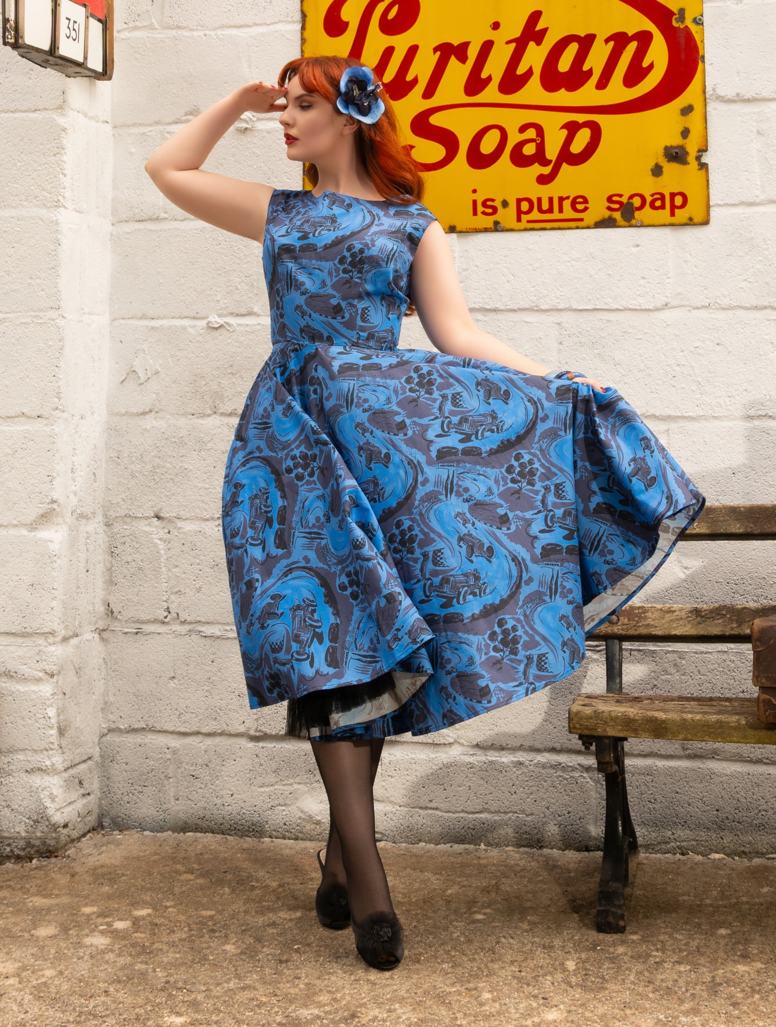 A woman with red hair wearing a blue and gray paisley-patterned dress, standing in front of a white brick wall with a vintage yellow 'Sutitan Soap' sign and a wooden bench in the background.