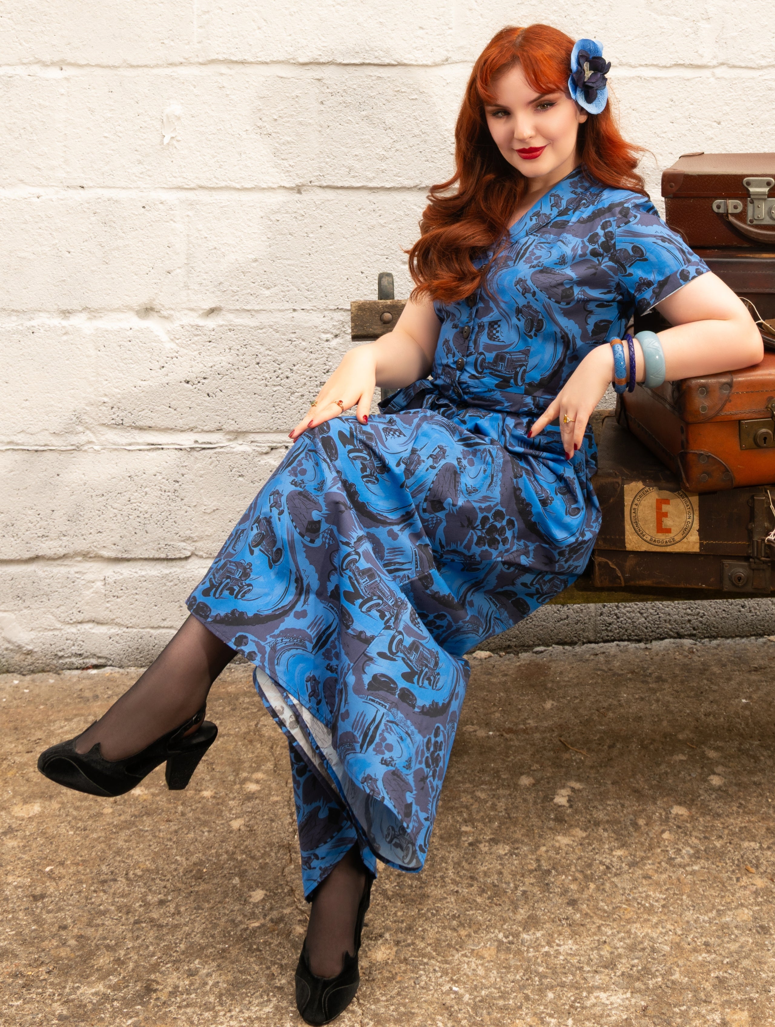 A woman with red hair sits on vintage brown suitcases against a white brick wall, wearing a blue and black patterned beach pyjamas with sheer black tights and black heels.