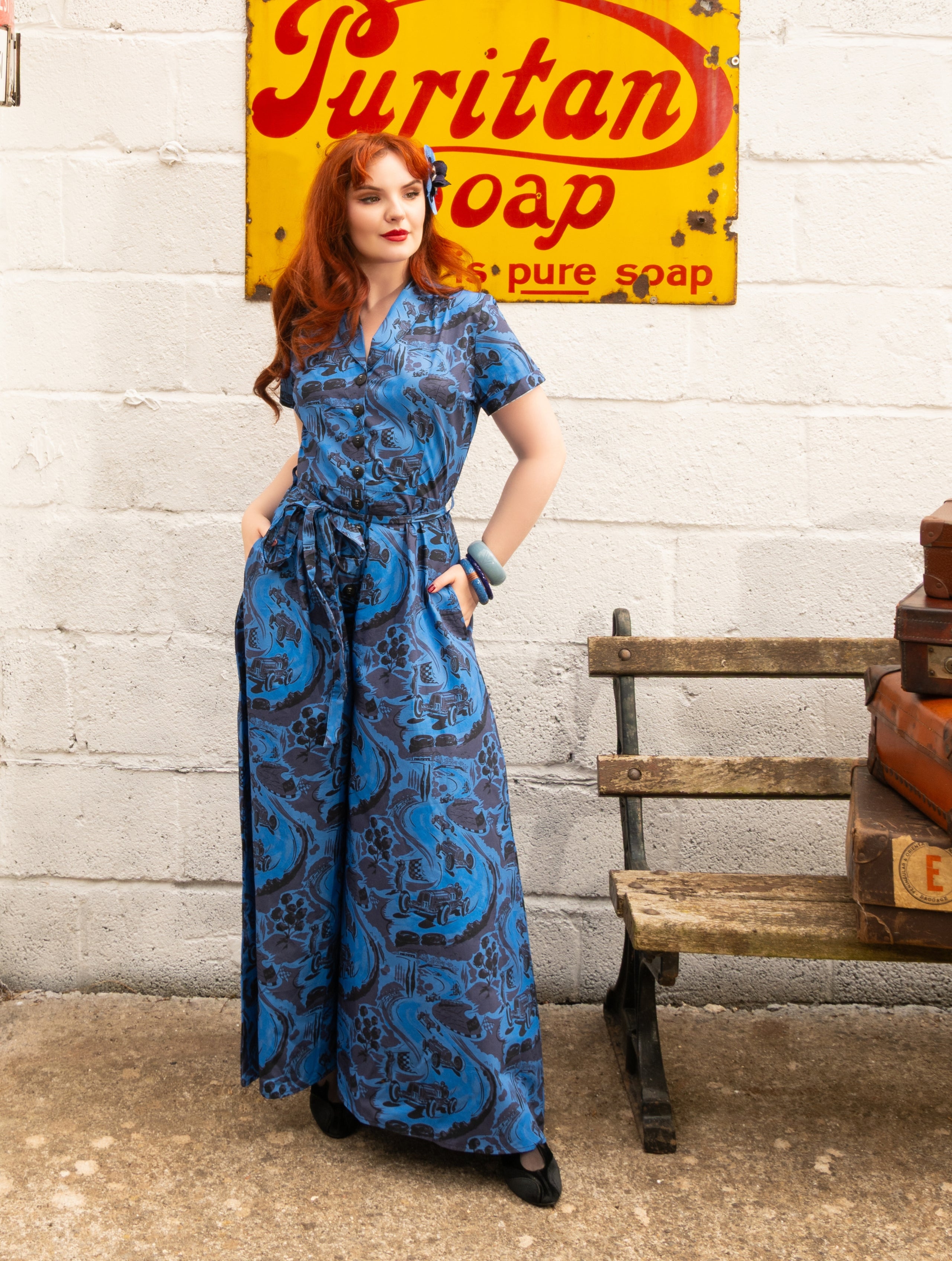 A woman with red hair models a blue and black patterned short-sleeve maxi dress with a tie waist, standing in front of a white brick wall with a vintage Puritan Soap sign and a wooden bench.