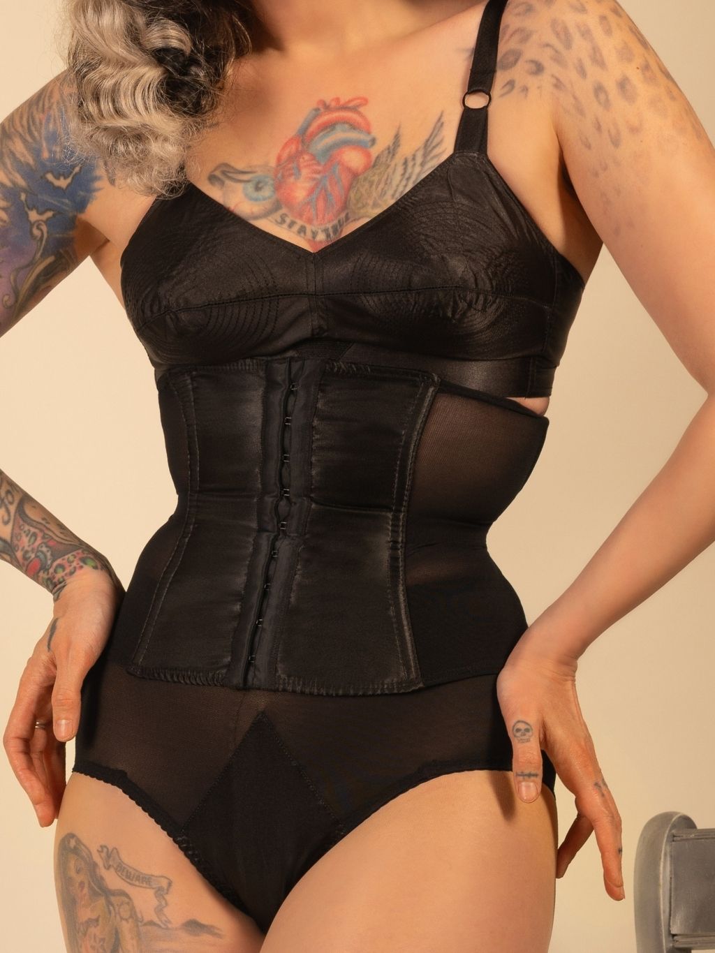 Person wearing a black  waspie corset and bodysuit with tattoos visible on arms and chest.