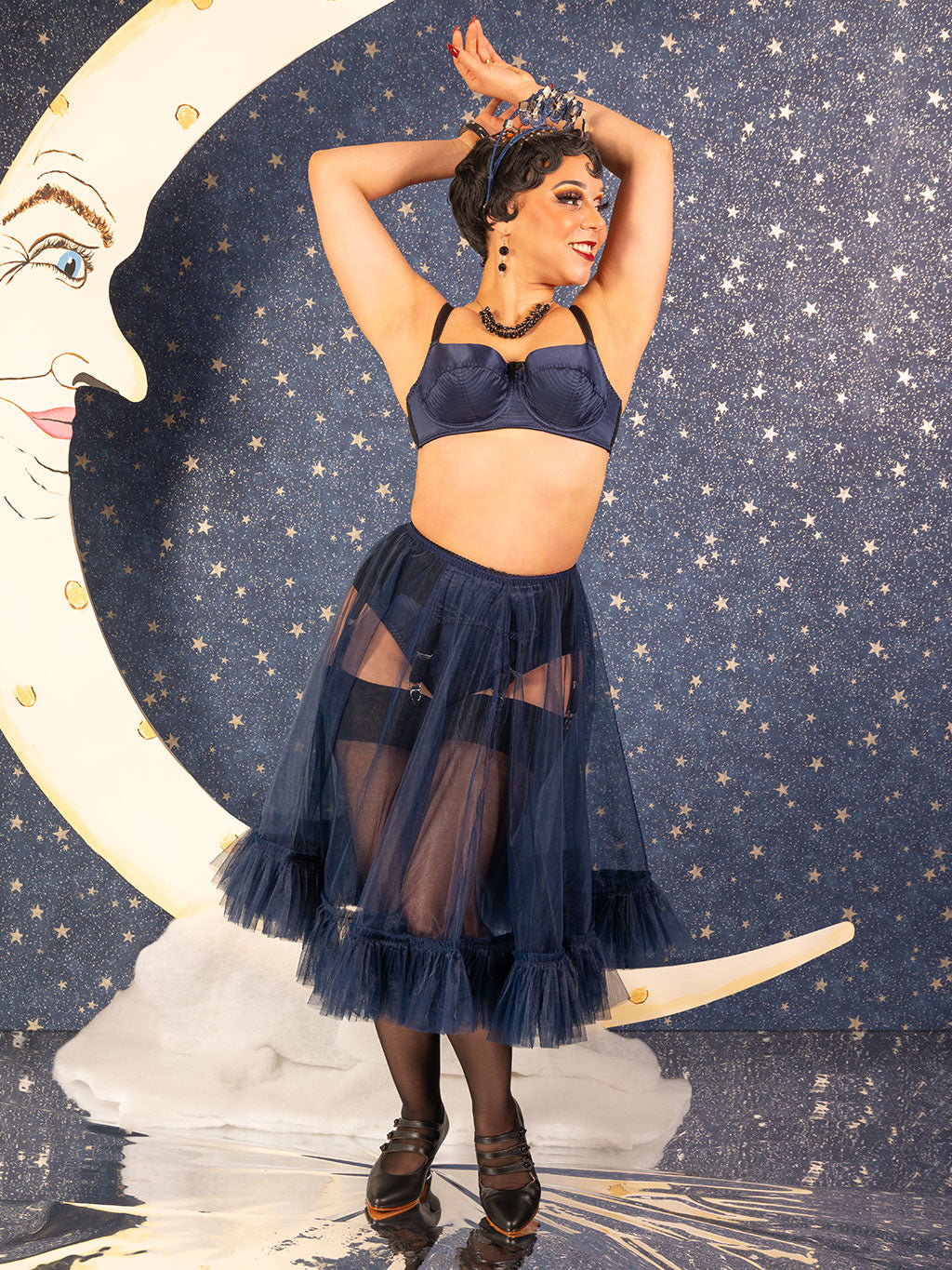 Woman in a vintage navy blue frilly petticoat and lingerie in front of a moon and star background with a starry background and crescent moon.