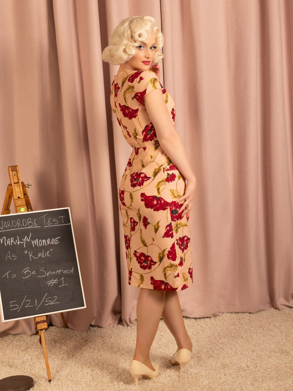 Woman in a vintage floral dress standing in front of a pink curtain with a blackboard sign.