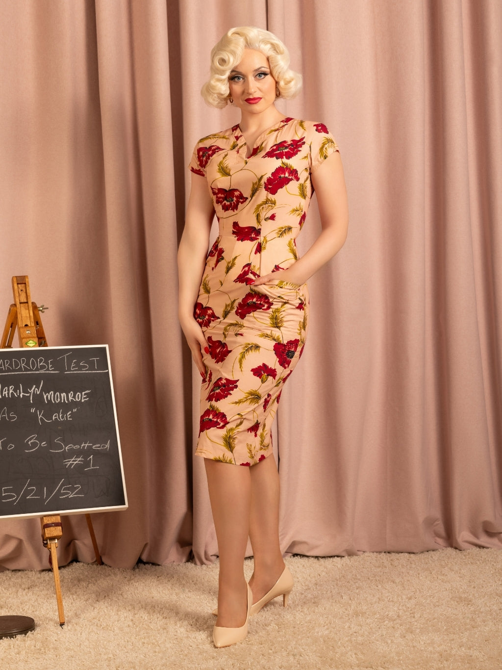 Woman in a vintage-style floral dress standing in front of a pink curtain with a blackboard in the background.