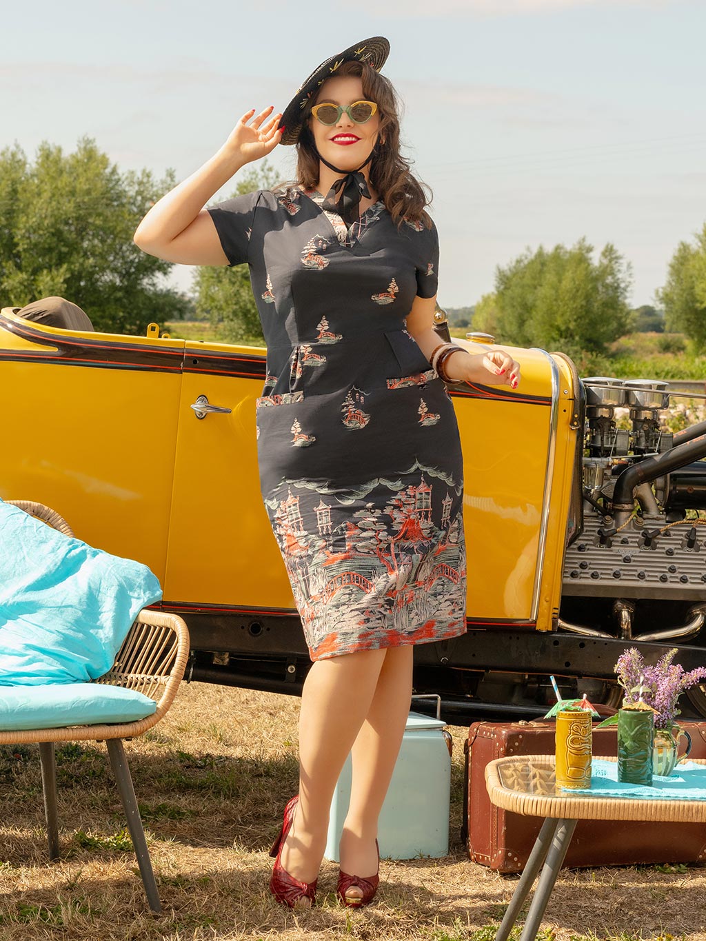 Woman in a vintage dress standing next to a yellow car with a scenic background