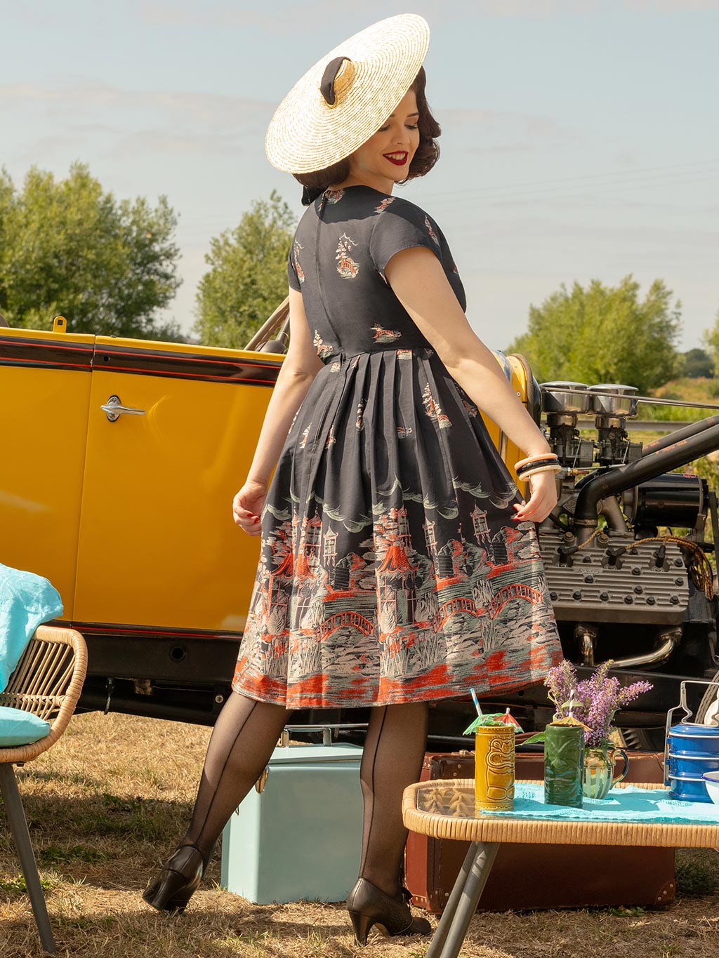 Woman in a vintage dress standing outdoors with a yellow car and picnic setup in the background