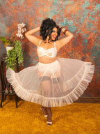 A woman modeling a vintage peach lingerie set consisting of a bra, matching garter belt, and a voluminous ruffled petticoat, posed against a textured orange and teal wall with a yellow rug.