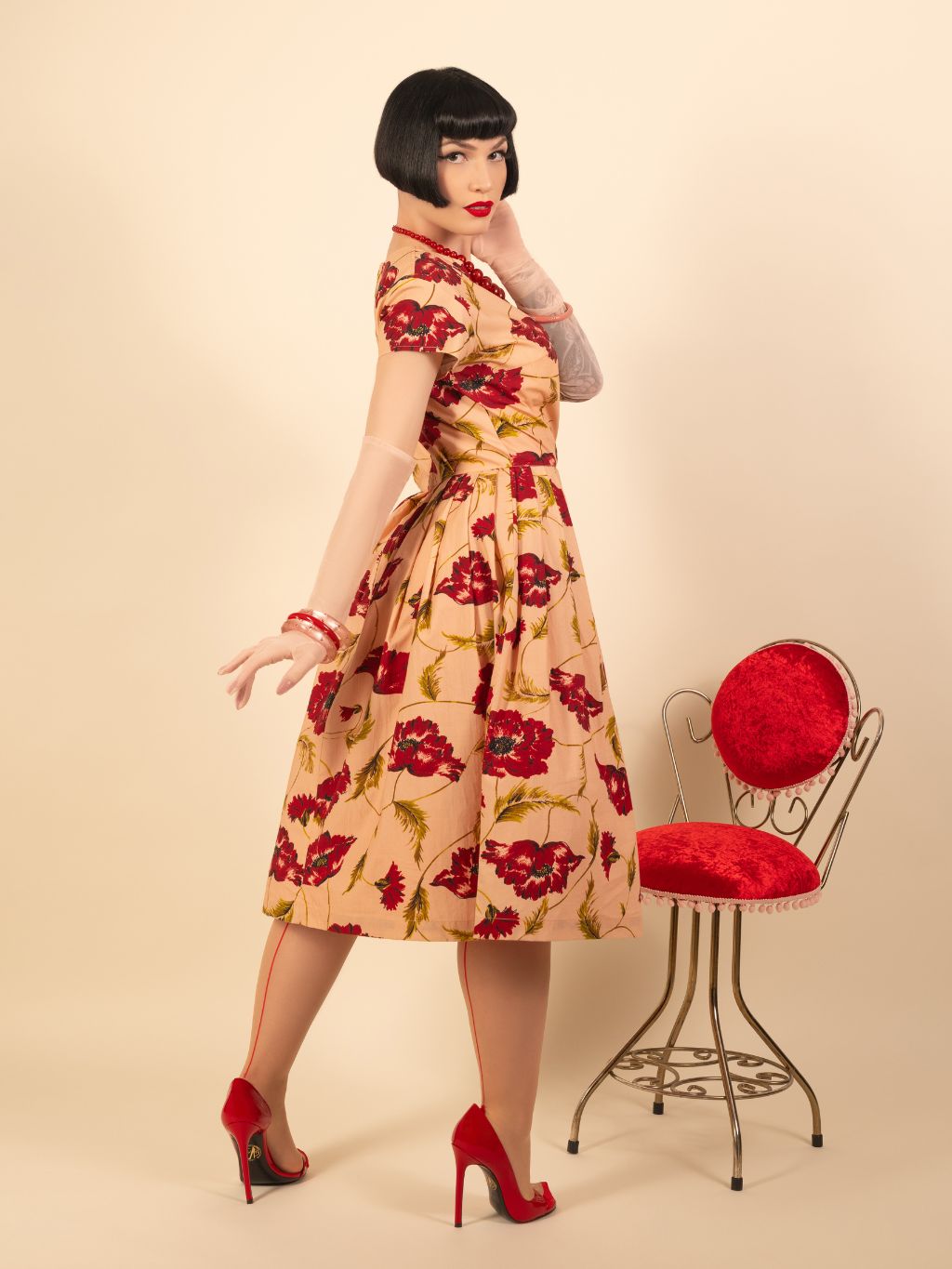 Woman wearing a floral dress standing next to a red cushioned chair on a beige background
