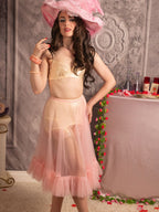 A woman models a light pink lingerie set consisting of a sheer bralette and a ruffled tulle skirt, paired with a large pink feather hat, in an ornate room setting.