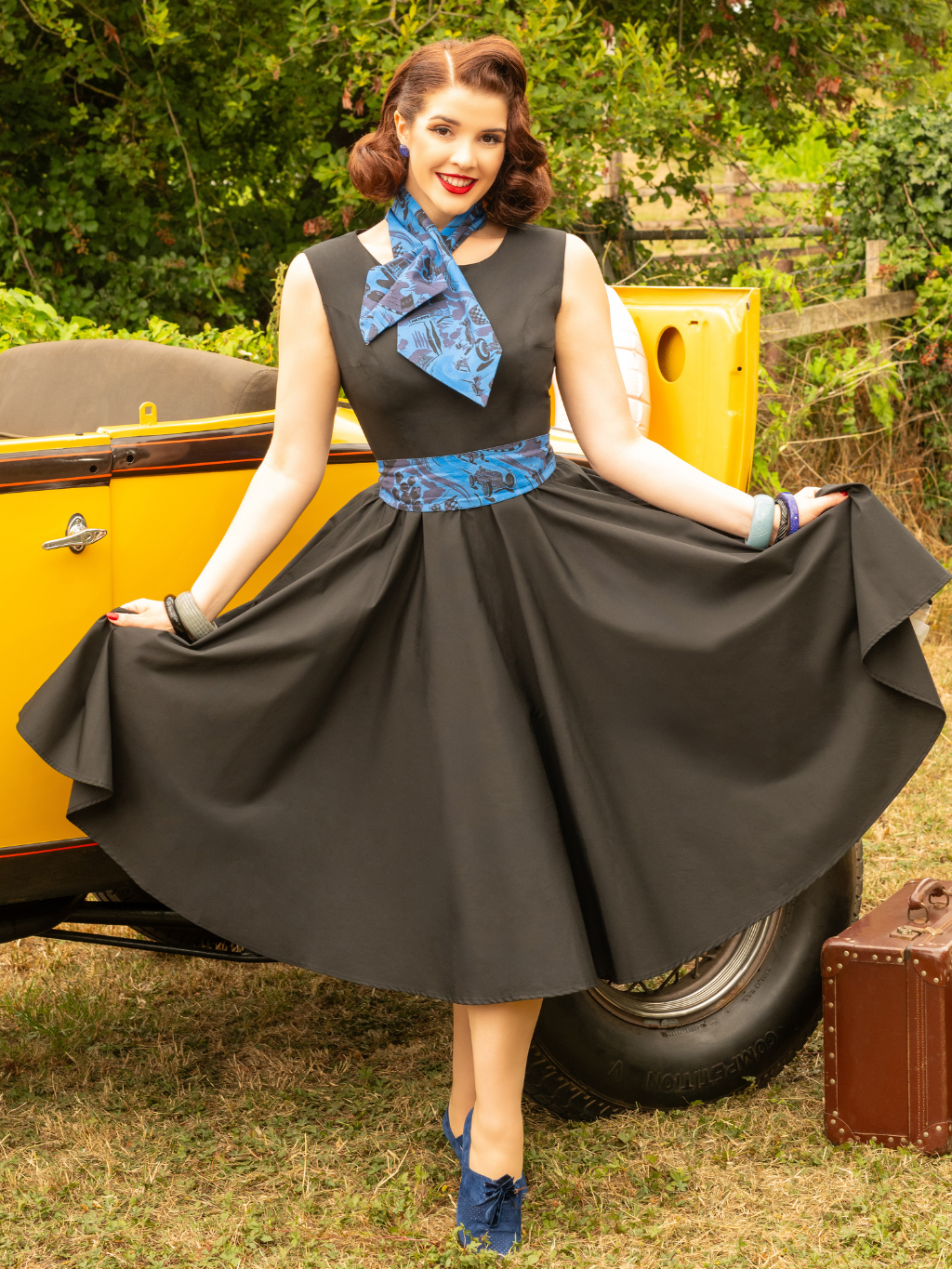 Woman in a vintage-style dress with a blue scarf, sitting in a yellow car with greenery in the background.
