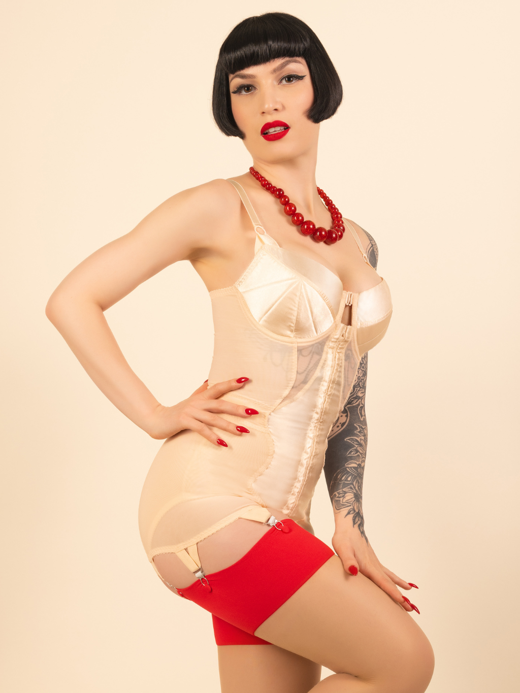 Woman wearing a 1950s peach corselette with red stockings against a beige background.