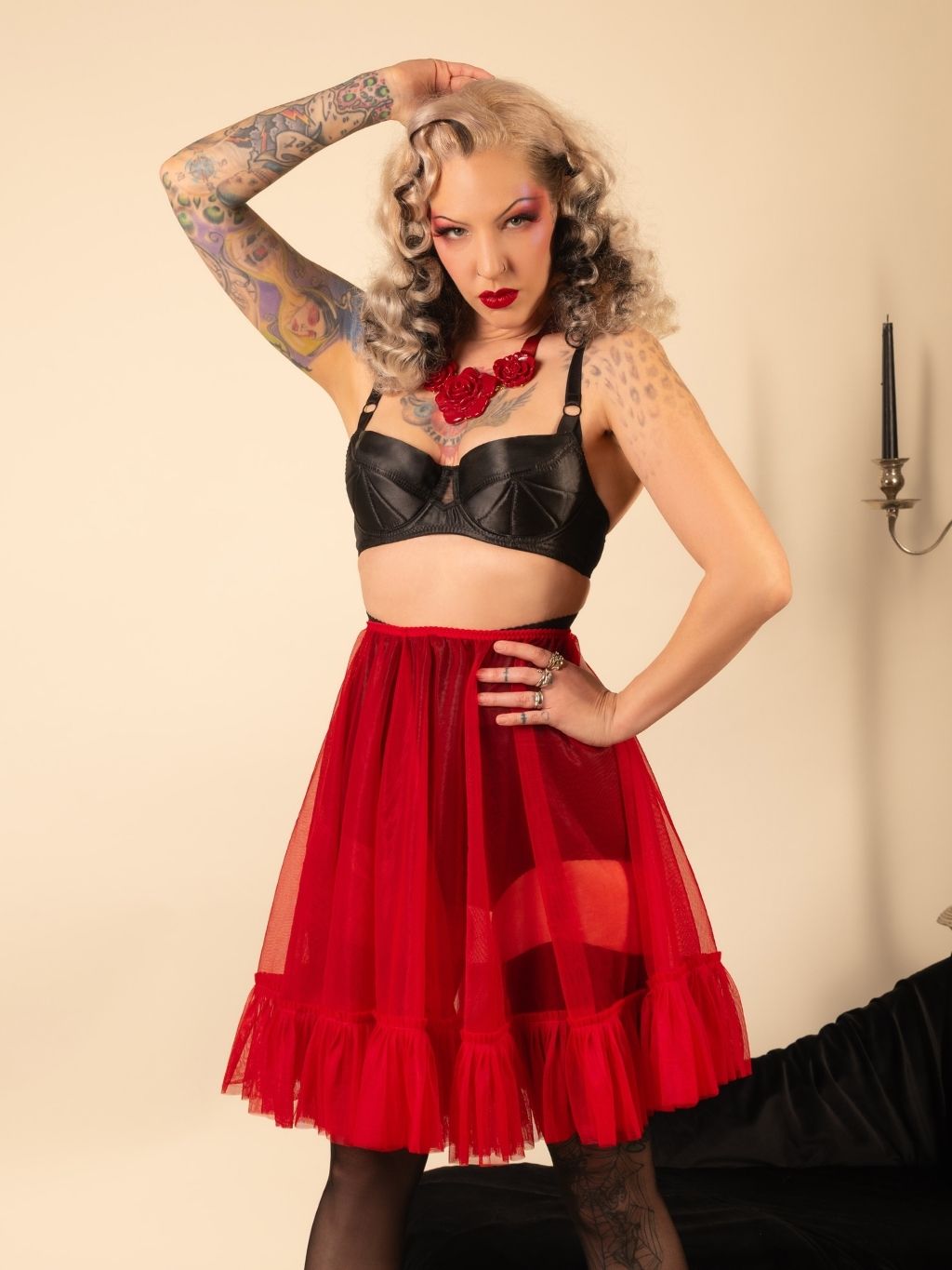 Woman wearing a black top and red frilly petticoat with tattoos and red makeup against a beige background