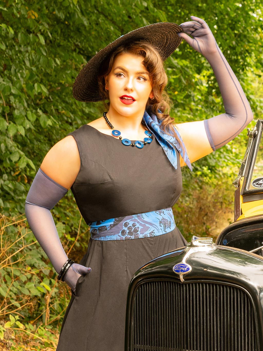 Woman in vintage attire standing next to an old car with a green leafy background