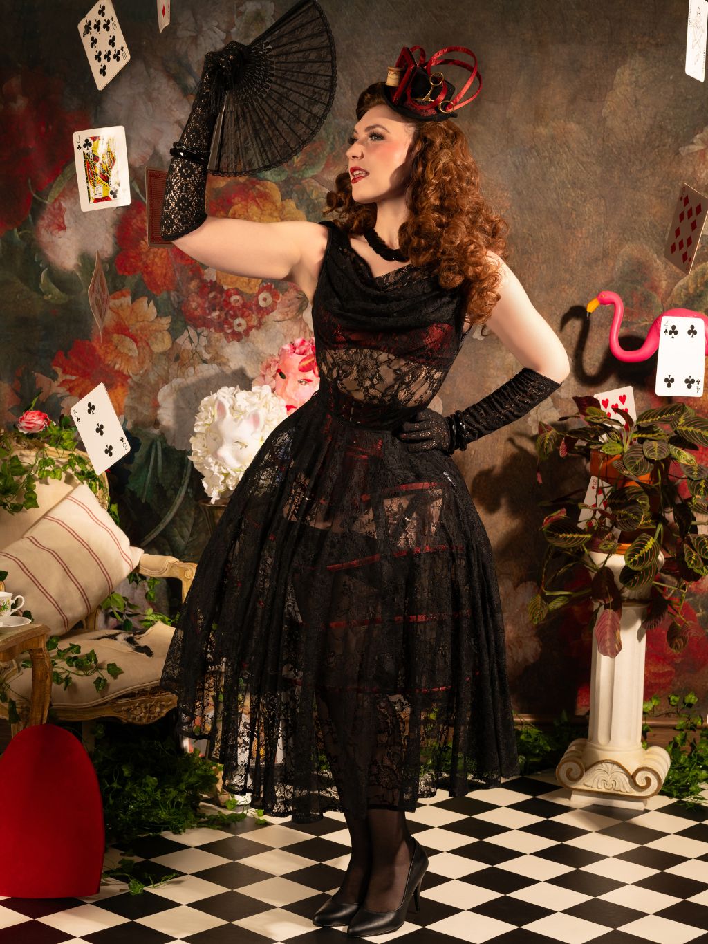 Woman in a black lace dress with a fan in a decorative setting with playing cards and flowers.