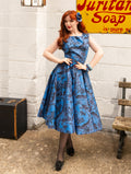 A woman stands in a vintage-style setting wearing a sleeveless blue and black patterned A-line dress with a fitted waist and flared skirt. She has red hair, black tights, and black shoes, posing in front of a white brick wall with a yellow sign and a wooden bench.