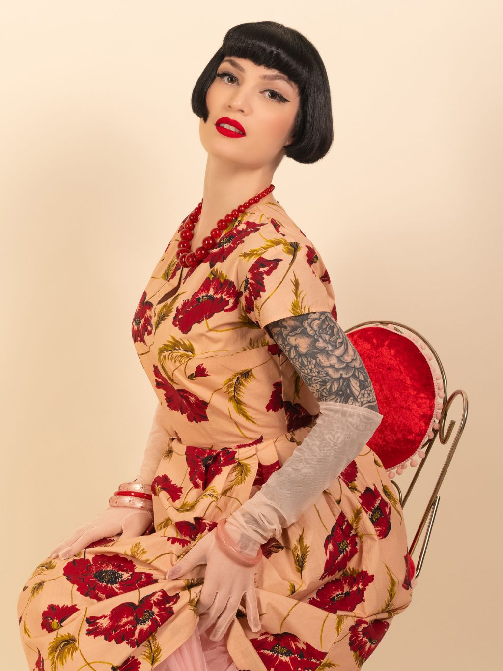 Woman in a floral dress with red flowers, and pink gloves, sitting on a chair against a beige background